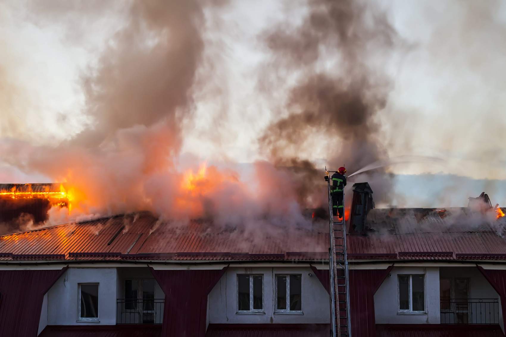 Burning fire flame with smoke on the apartment house roof in the city, firefighter on the ladder extinguishes fire.