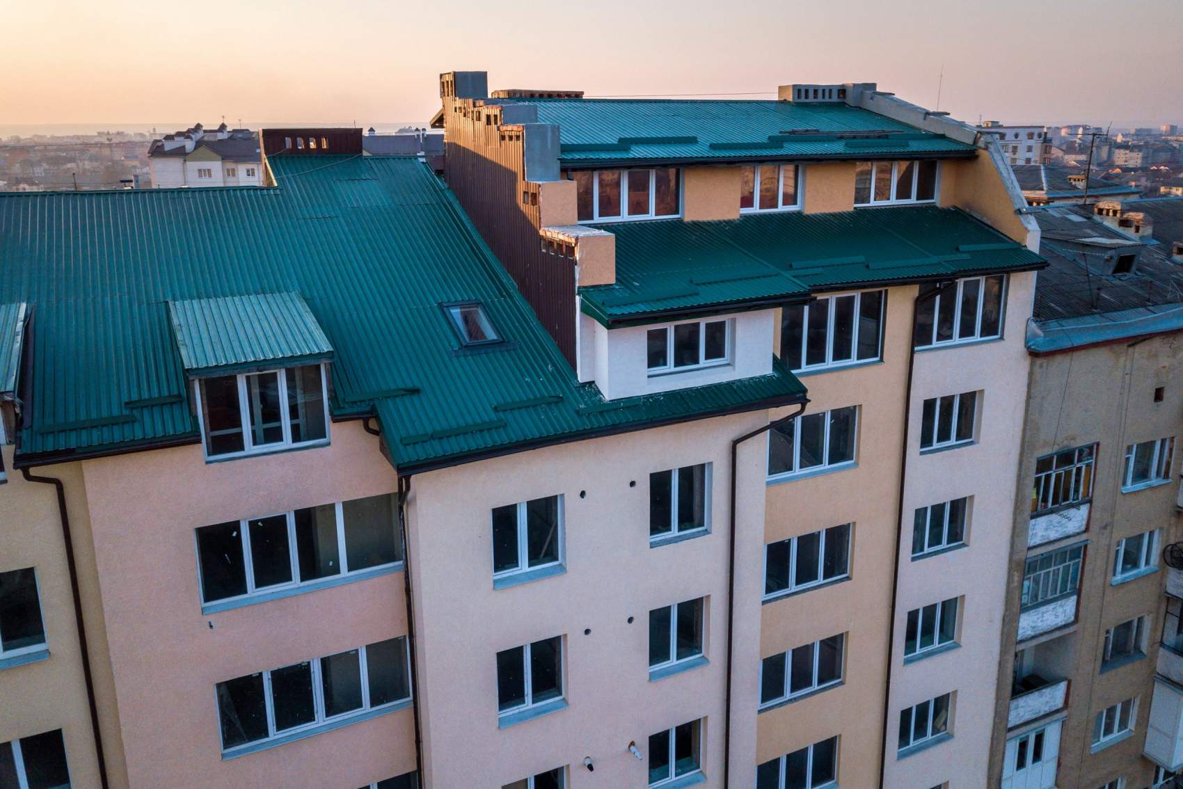 Aerial view of attic annex room exterior with plastic windows, roof and walls covered with green metal siding planks, new gutter system on top of high multi-storey apartment building.