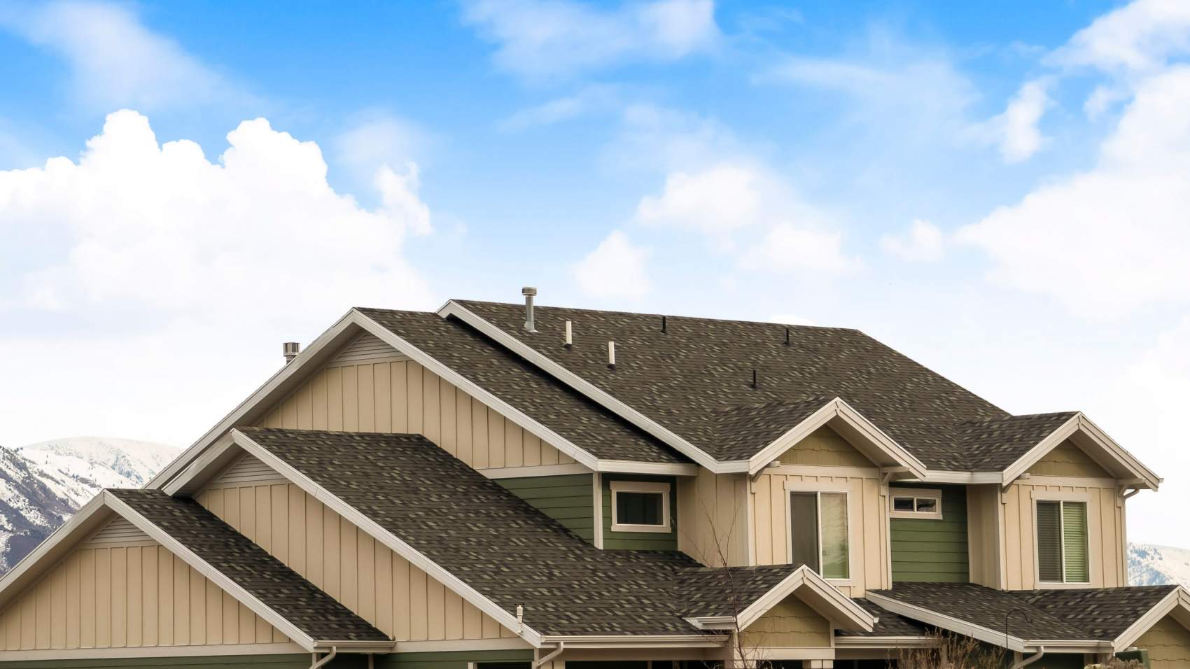 Panorama frame House exterior with view of the dark pitched roof against a cloudy blue sky. A mountain blanketed with snow can be seen behind the home.
